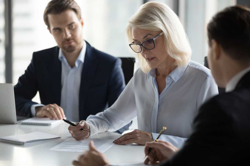 Businessmen sitting at desk headed by middle aged serious concentrated female in eyeglasses checking agreement document before signing it. Financial director ready affirm official paper with signature