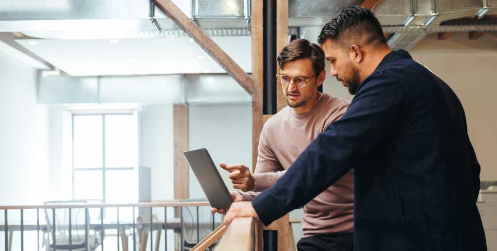 Business man discussing a tech project with his colleague in an office. Two business men using a laptop on an interior balcony. Male professionals working together.